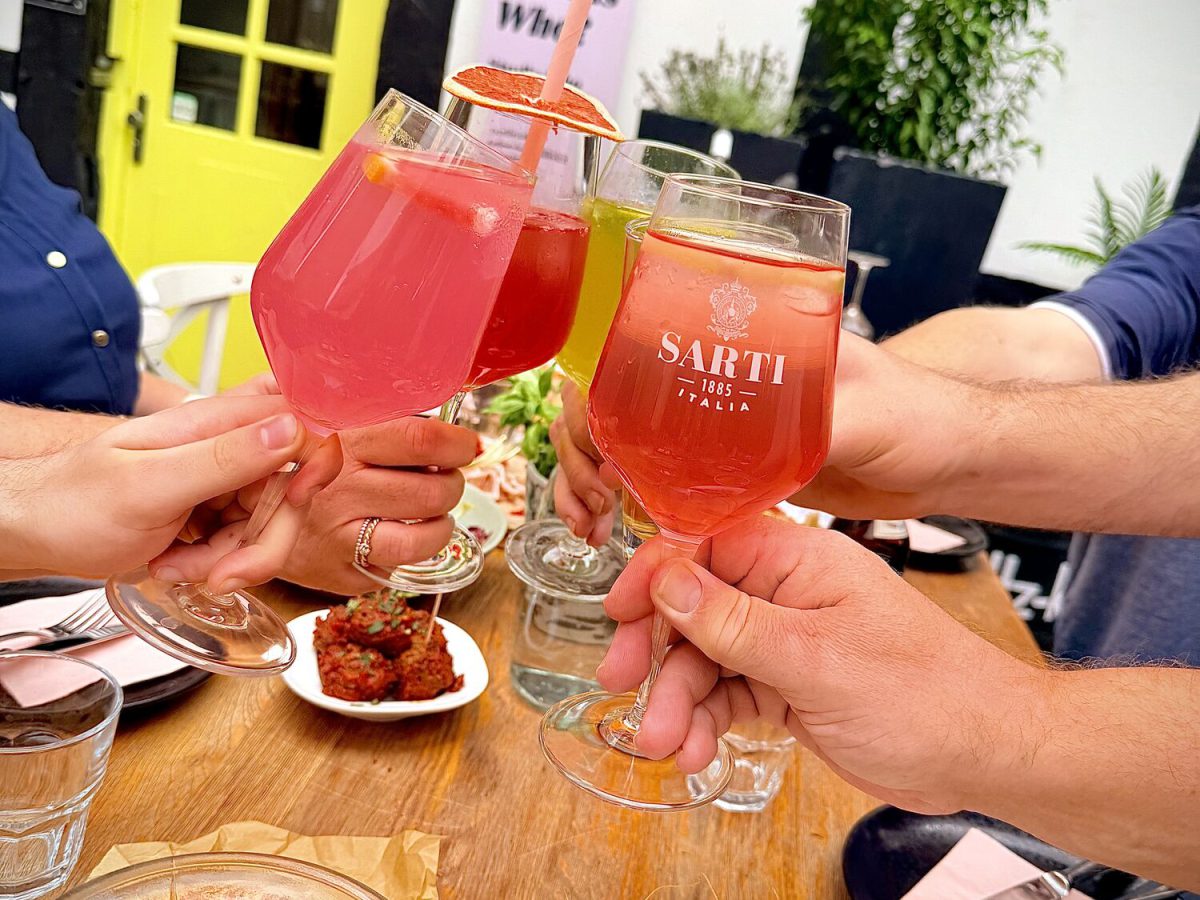 Zwei Gäste stoßen mit SARTI Spritz vor der gelben Tür des Studio Who an