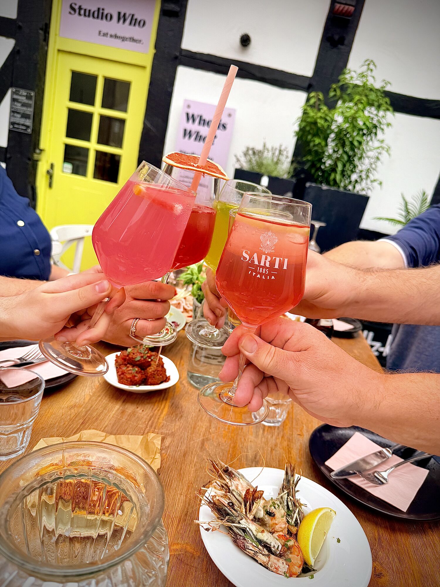 Zwei Gäste stoßen mit SARTI Spritz vor der gelben Tür des Studio Who an