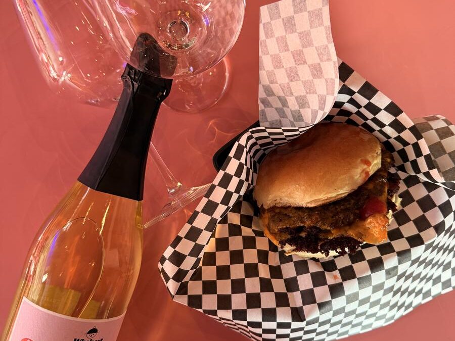 Draufsicht auf einen Burger in Karopapier neben zwei Gläsern und einer Flasche Whobert Chardwhonnay Sekt auf pinkem Tisch