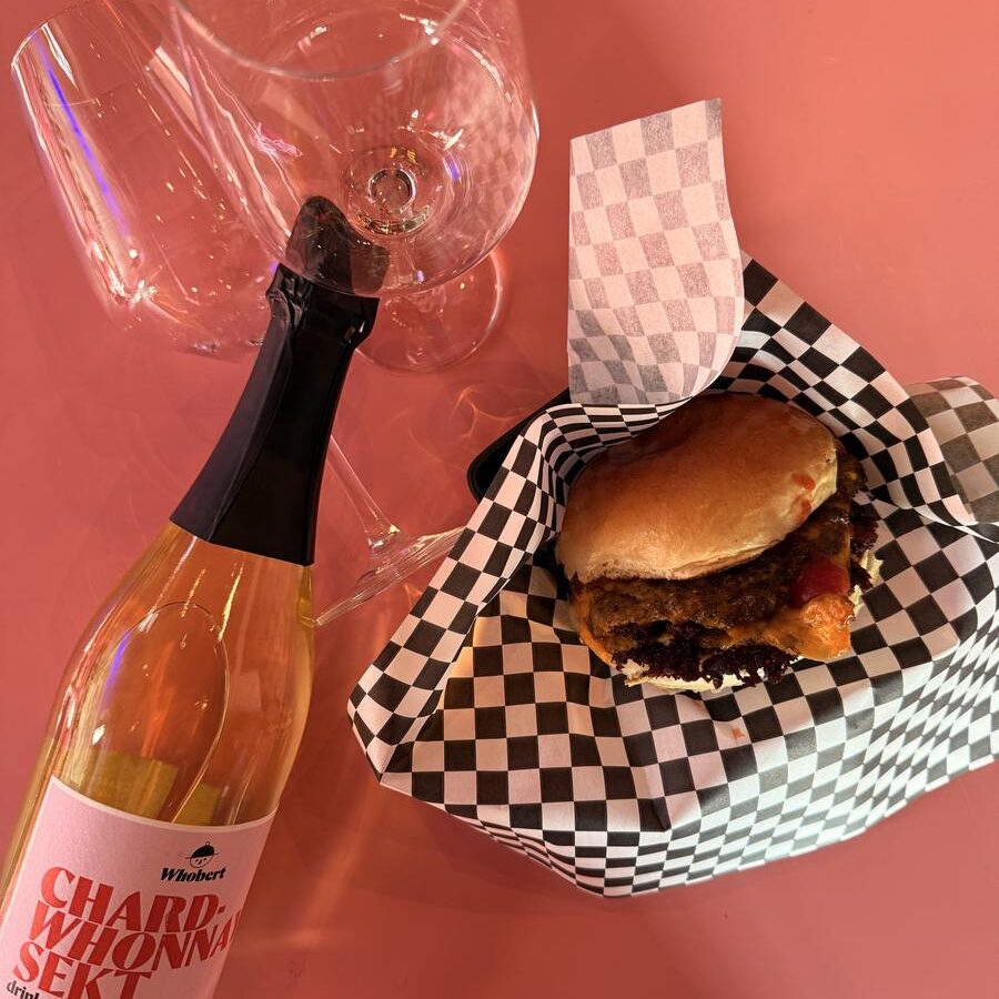 Draufsicht auf einen Burger in Karopapier neben zwei Gläsern und einer Flasche Whobert Chardwhonnay Sekt auf pinkem Tisch