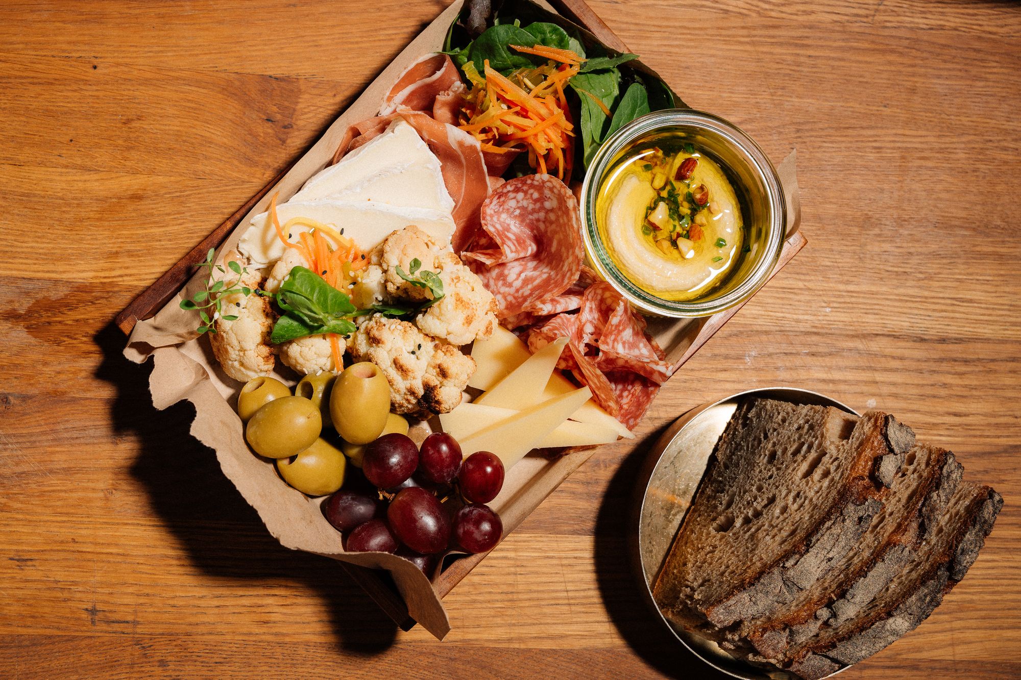 Daily-Who-Box mit Schinken, Käse, Blumenkohl, Oliven und Hummus, daneben Sauerteigbrot