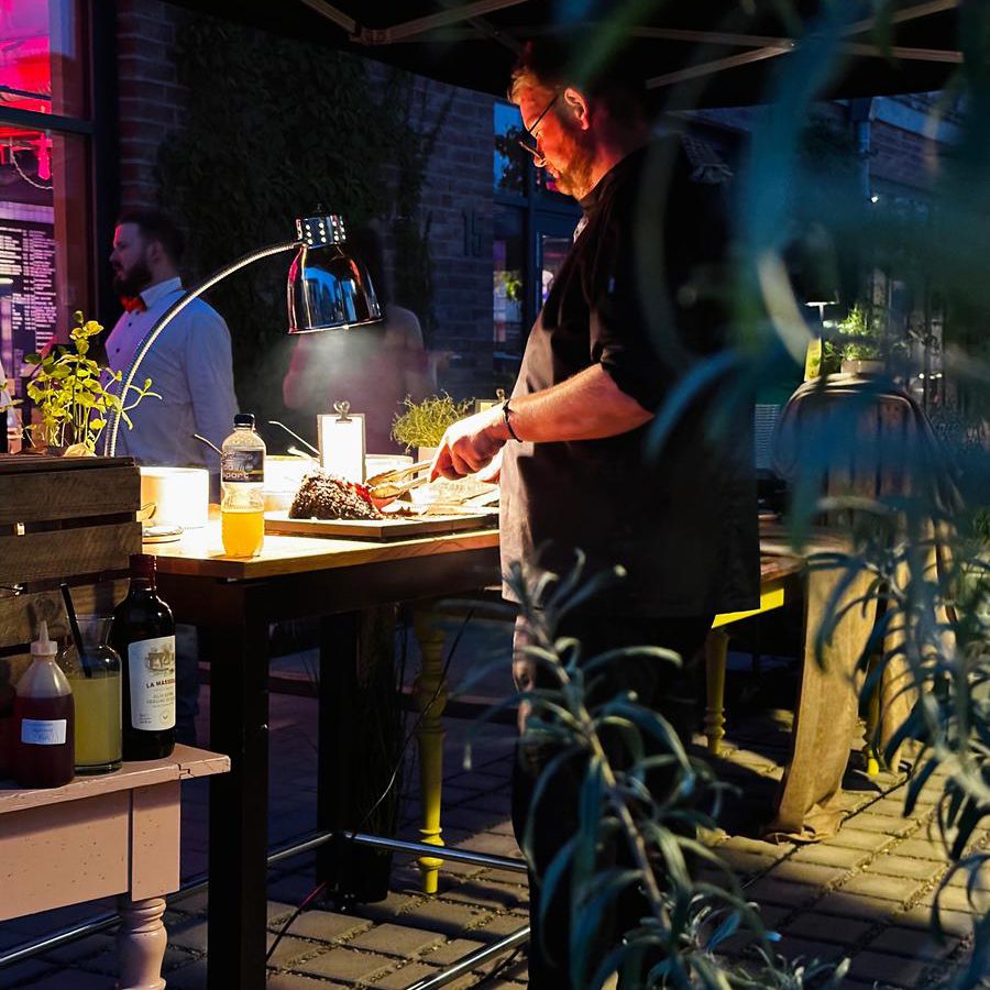 Koch grillt abends bei einer Hochzeitsfeier im Freien, im Hintergrund Gäste