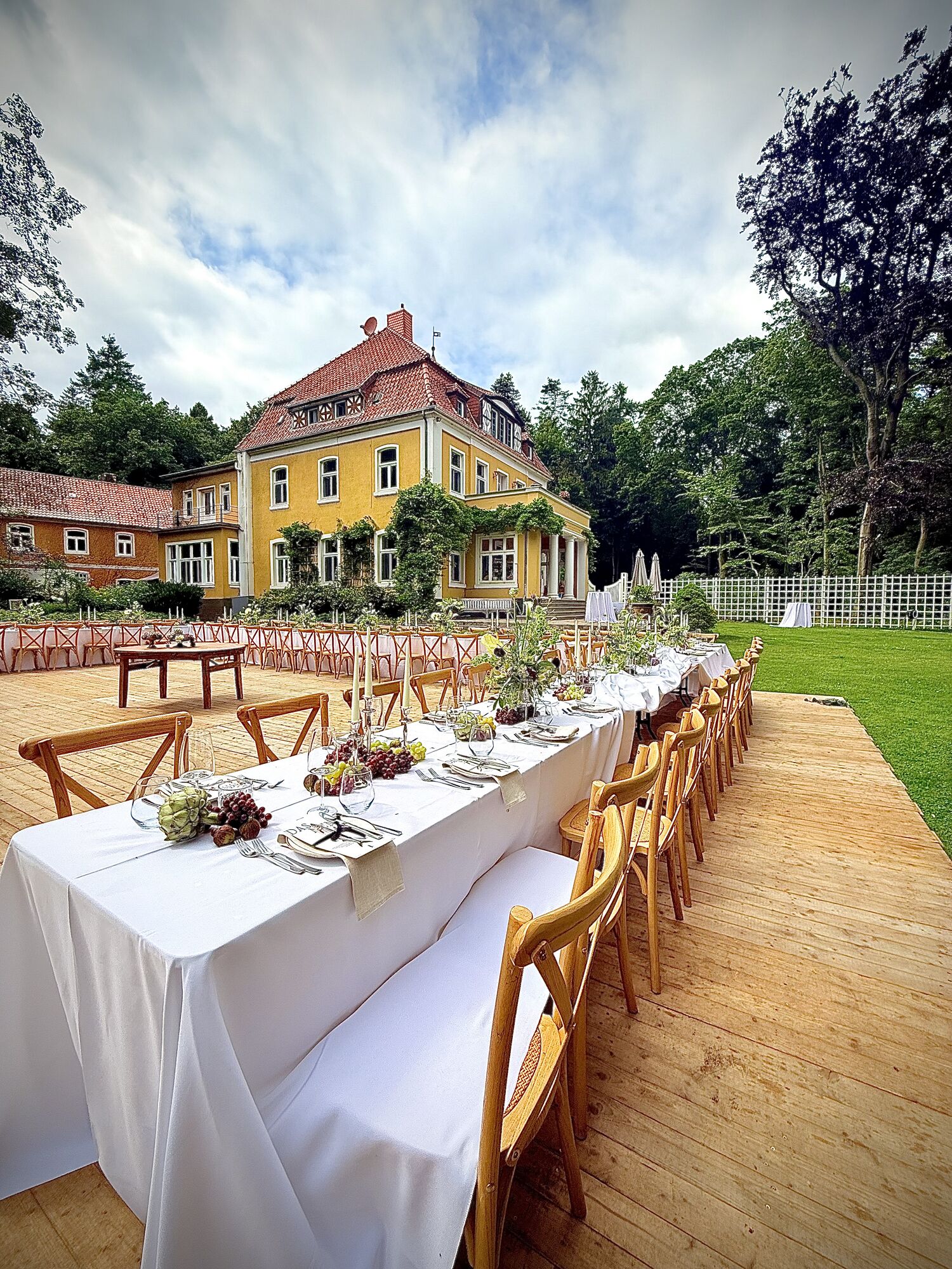 Lange weiße Hochzeitstafel mit Holzstühlen im Garten einer Gutsvilla