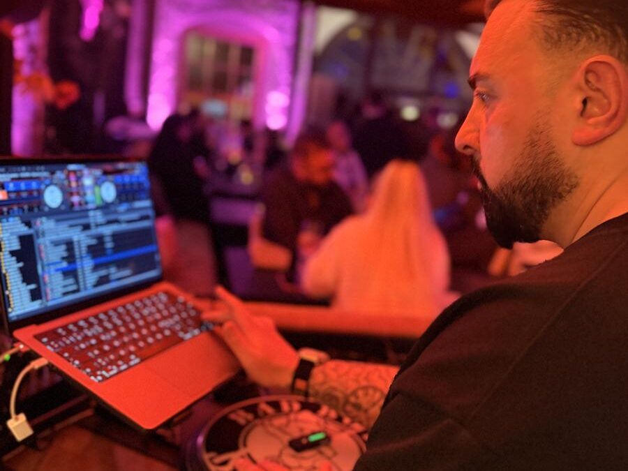 DJ Shain steht an Laptop und Plattenteller bei einem Abendevent, im Hintergrund tanzen Gäste in pink-violettem Licht
