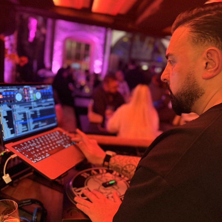 DJ Shain steht an Laptop und Plattenteller bei einem Abendevent, im Hintergrund tanzen Gäste in pink-violettem Licht